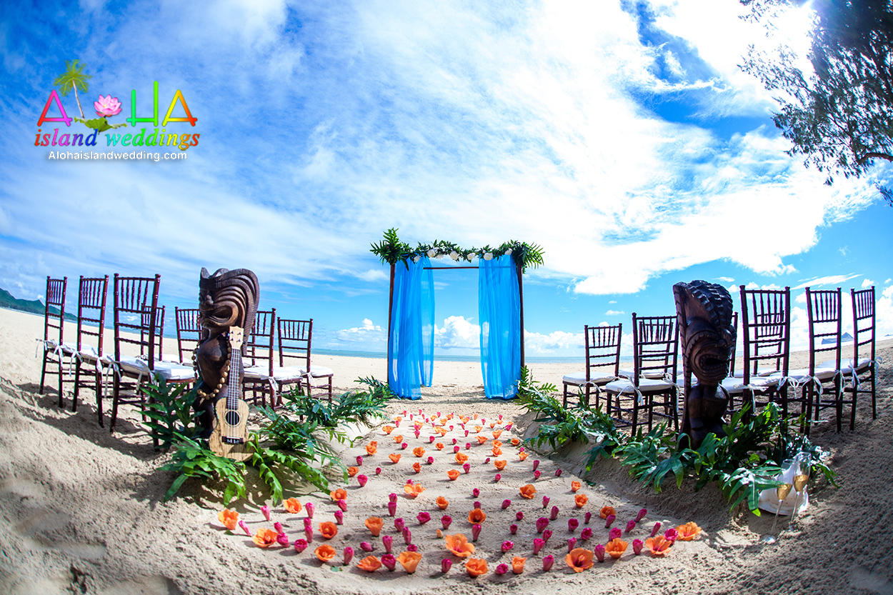 Blue and white theme with wooden chiavari chairs and Hawaiian tiki gods stand guard with dark pink and orange hibiscus for the aisle way